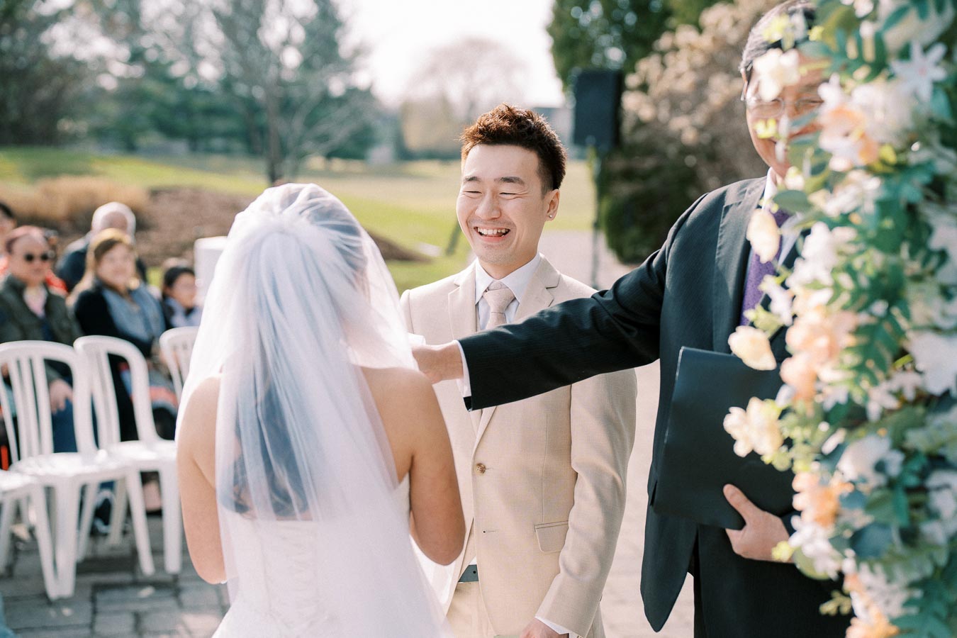 Outdoor wedding ceremony with a bride in a white dress and veil standing opposite a smiling groom in a light suit, surrounded by guests and floral decorations.