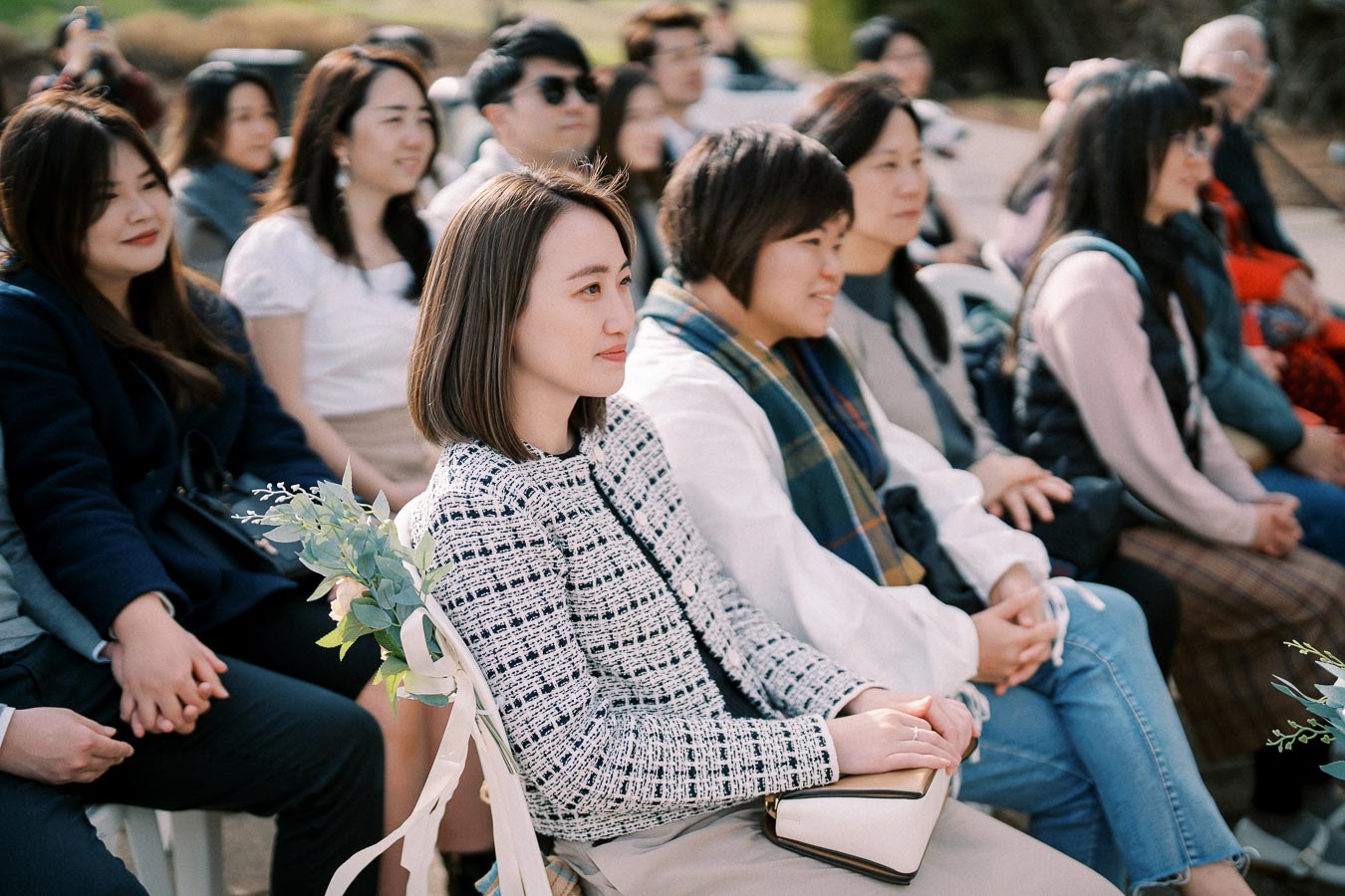 A group of people seated outdoors, attentively watching an event. The focus is on a woman in a patterned jacket holding a bouquet, surrounded by others engaged in the moment. Ideal for depicting audience engagement at a formal ceremony or gathering.