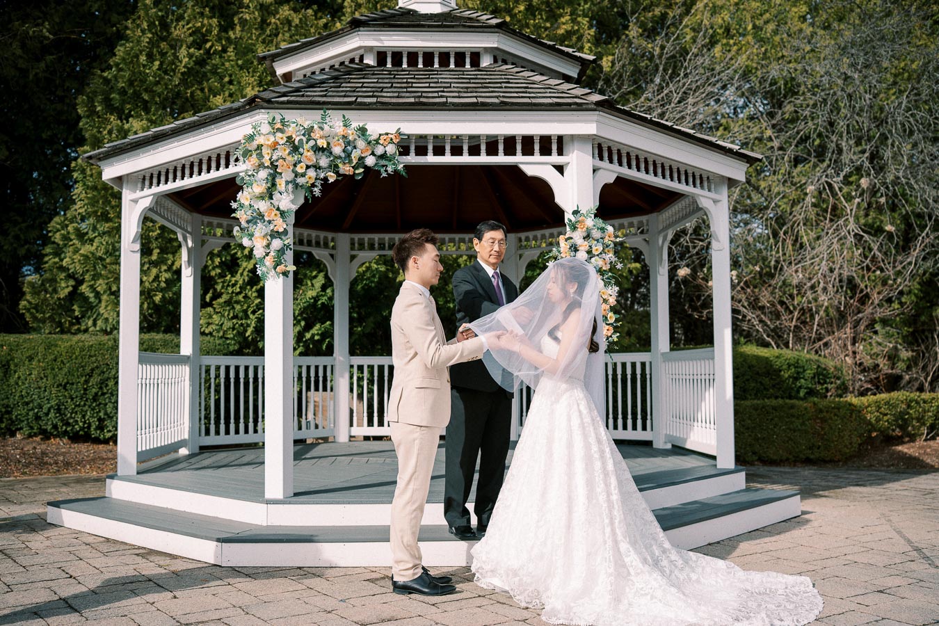 Wedding ceremony in an outdoor gazebo surrounded by greenery, featuring a bride in a lace gown and veil holding hands with the groom in a beige suit, as an officiant stands nearby.