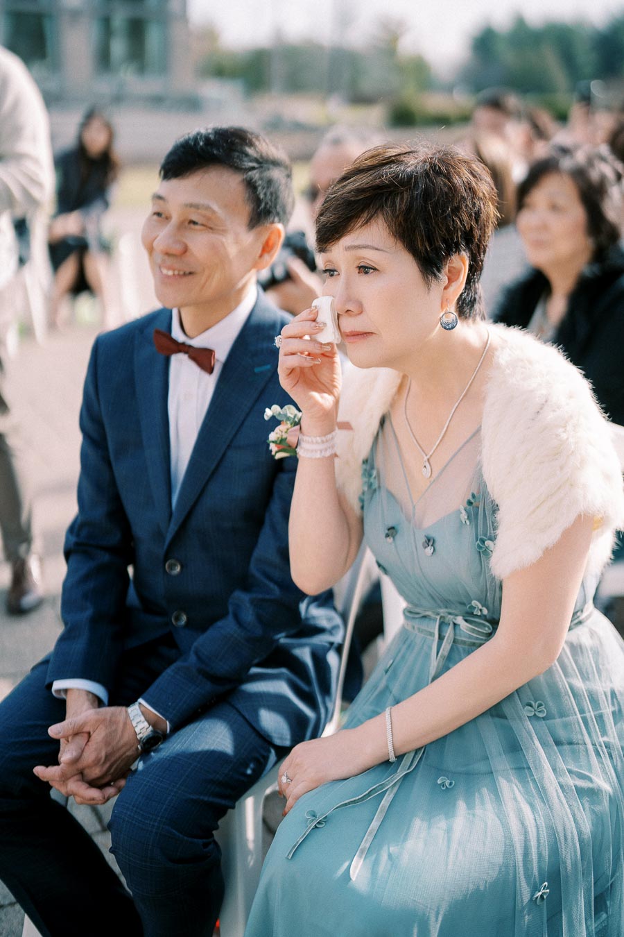 Elegant couple at an outdoor event, the woman in a blue dress with a fur stole wipes a tear, seated beside a smiling man in a blue suit with a red bow tie.