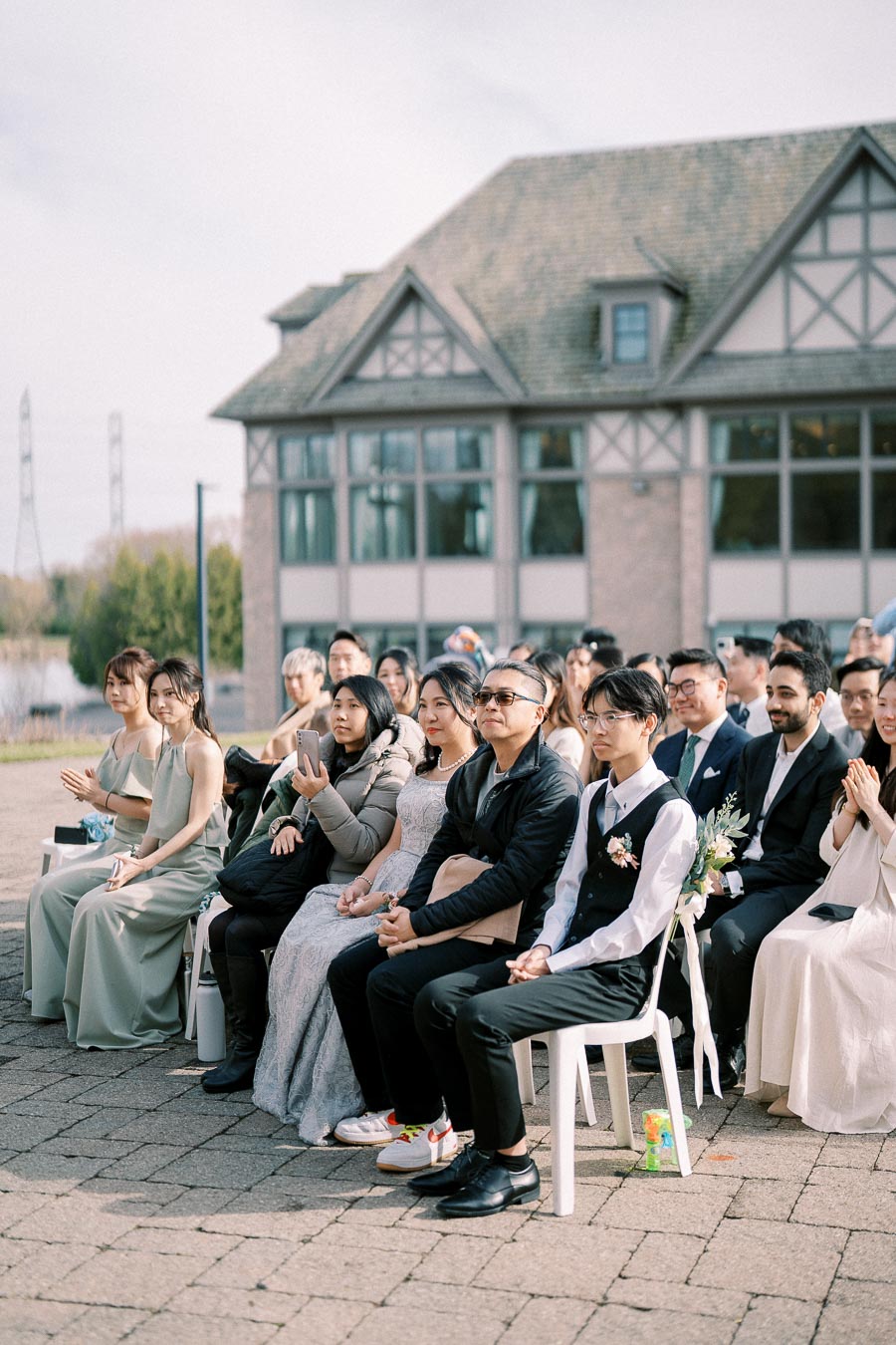 Guests seated at an outdoor wedding ceremony, attentively watching the event, with a charming, rustic building in the background.