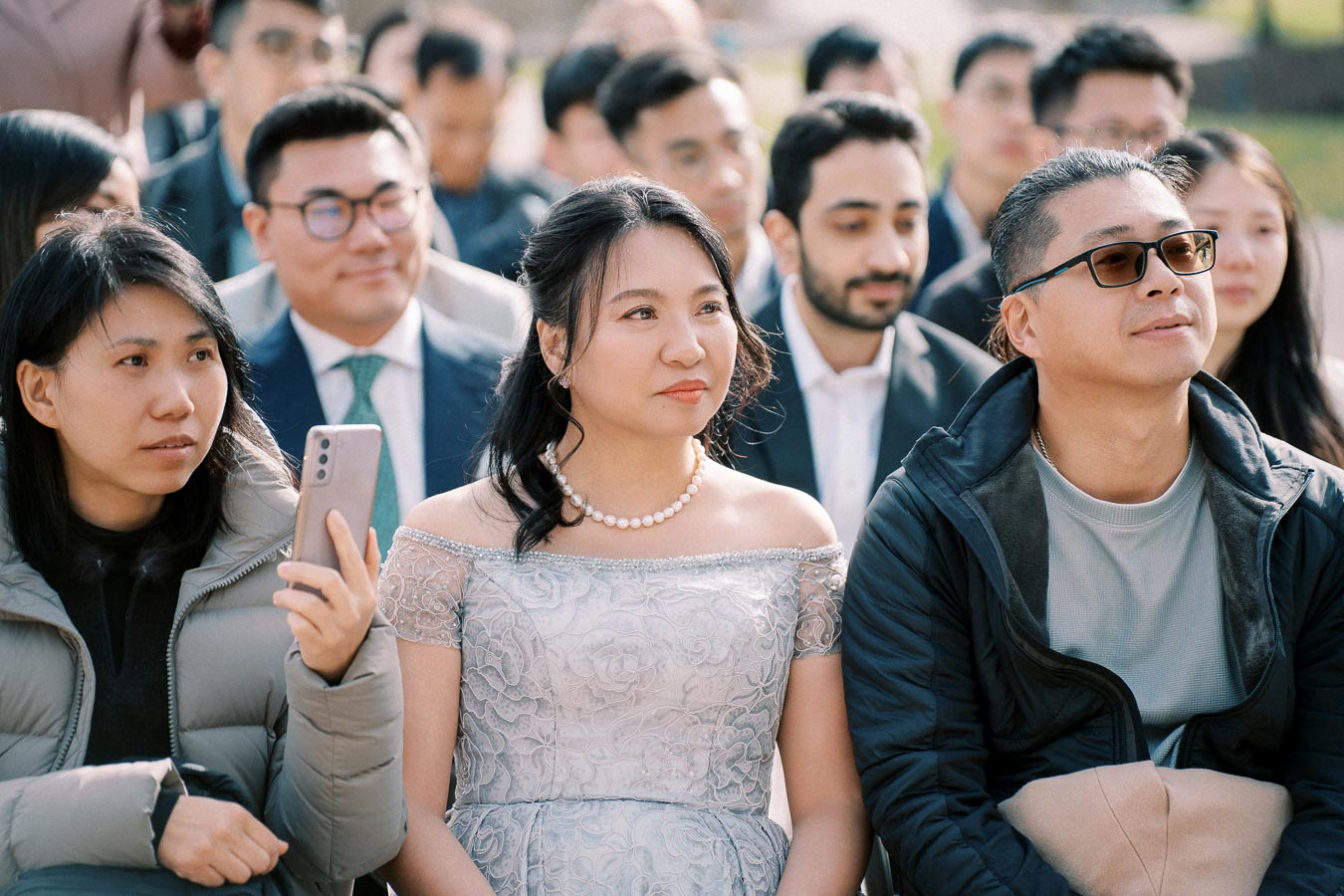 A diverse group of people seated outdoors, including a woman in a lace dress and pearl necklace, attentively watching an event, with some holding smartphones.