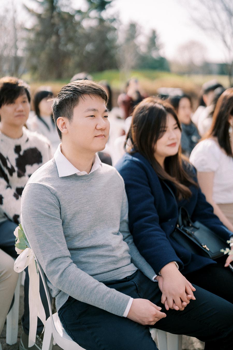 A couple seated outdoors holding hands, surrounded by other people attending an event, with a scenic background of trees and a sunny day.
