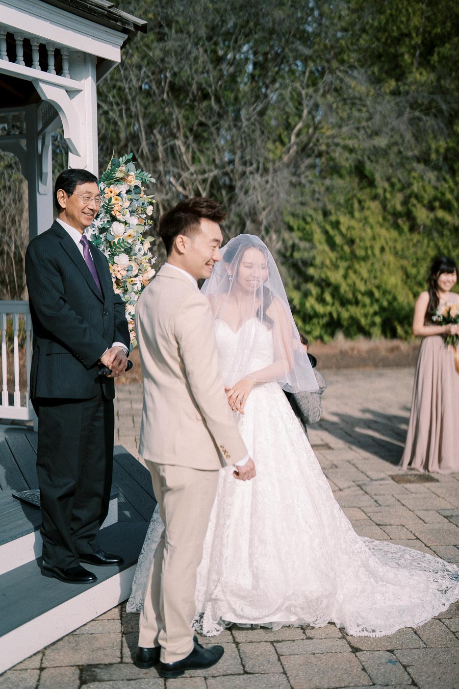 Outdoor wedding ceremony with a bride in a lace gown and veil smiling beside the groom in a beige suit, standing near a floral arch.