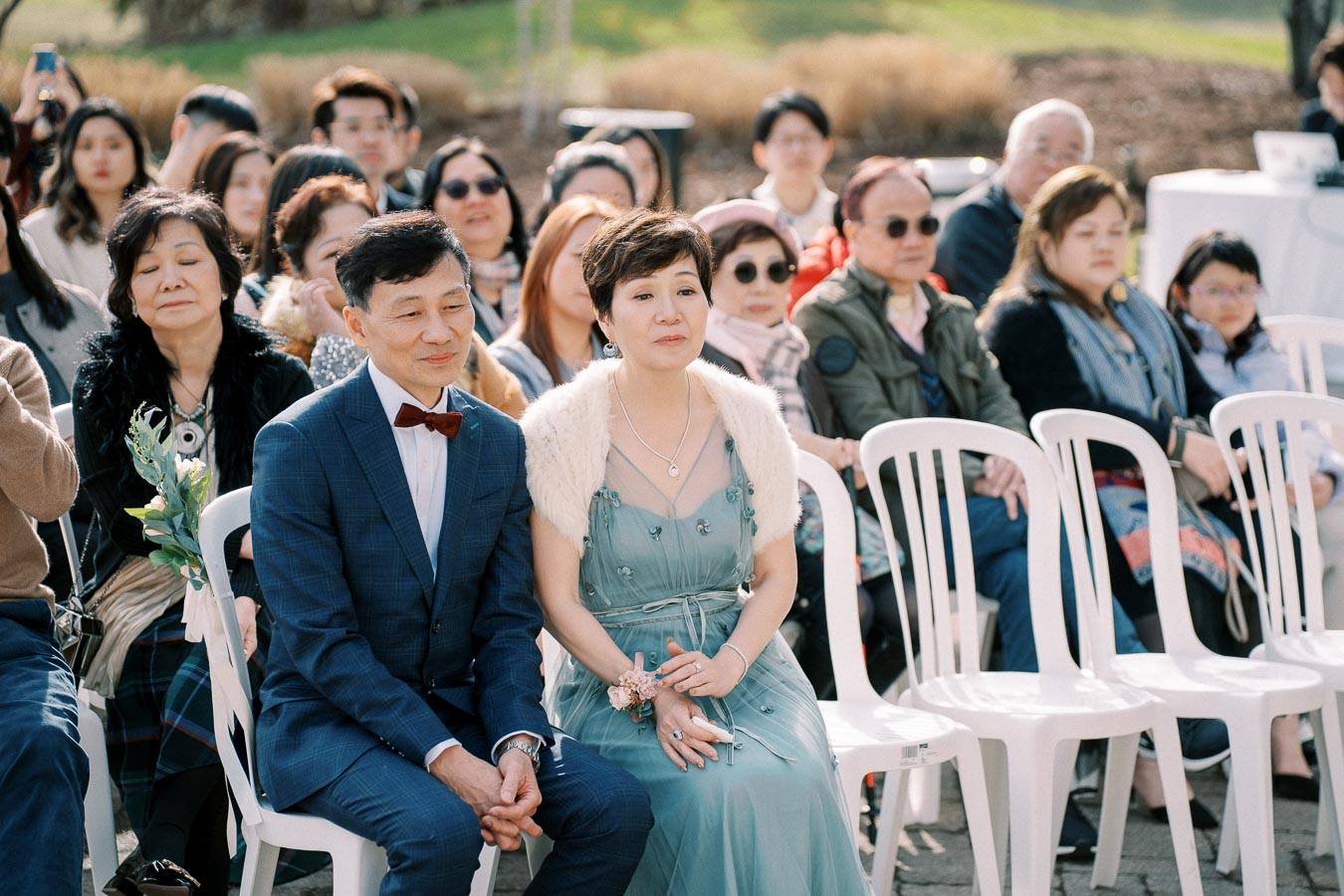 Outdoor wedding ceremony with elegantly dressed guests seated, featuring a couple in the front row, the man in a blue suit and bow tie, and the woman in a teal gown with a fur shawl, both looking content and engaged.