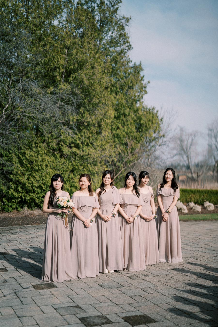 Outdoor wedding portrait featuring six bridesmaids in matching taupe dresses standing on a stone pathway with a background of lush green trees.