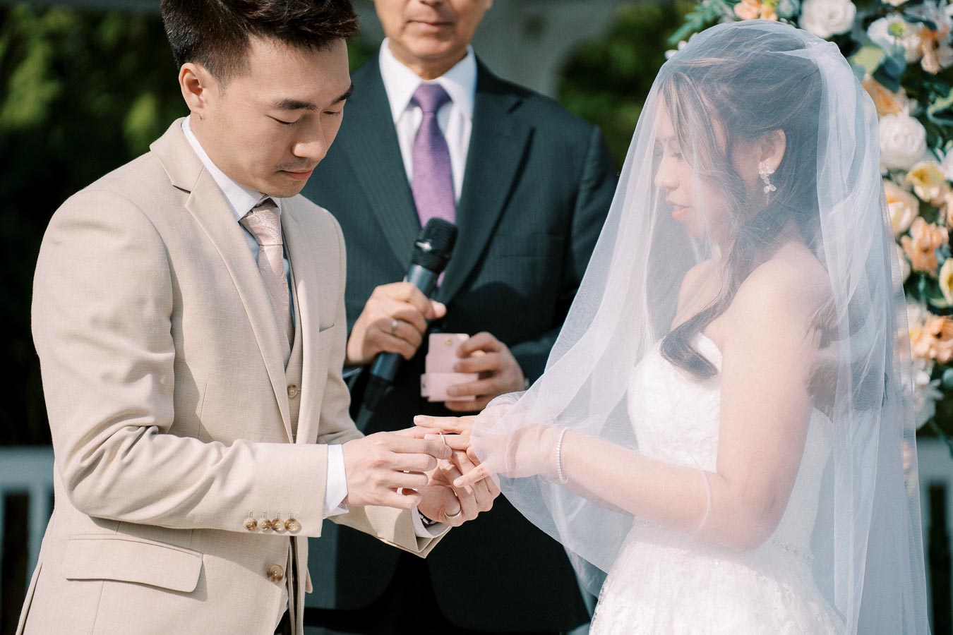 Bride and groom exchanging rings during an outdoor wedding ceremony, with a man officiating in the background, surrounded by floral decorations.