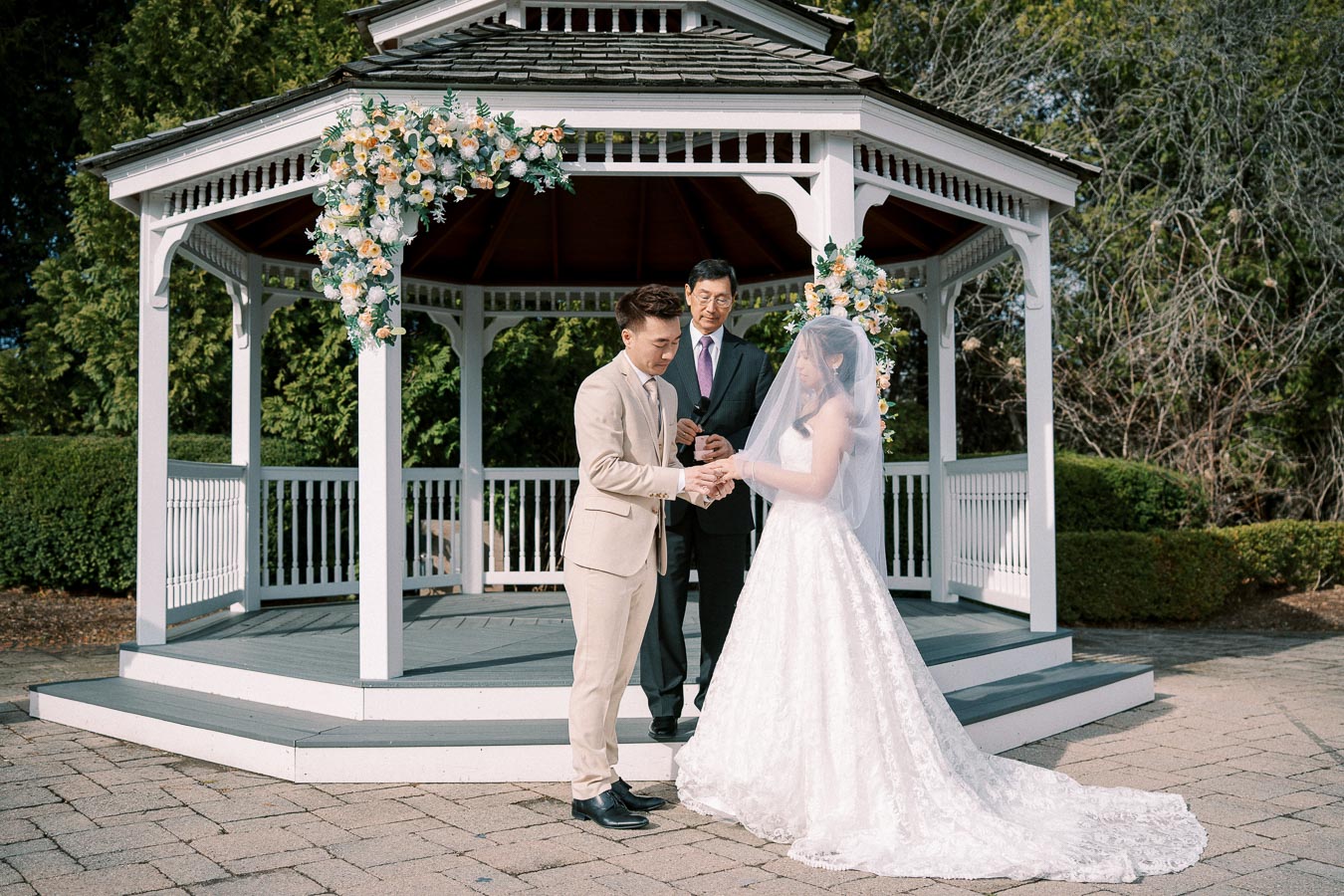 Bride and groom exchanging vows under a floral-decorated gazebo during an outdoor wedding ceremony.