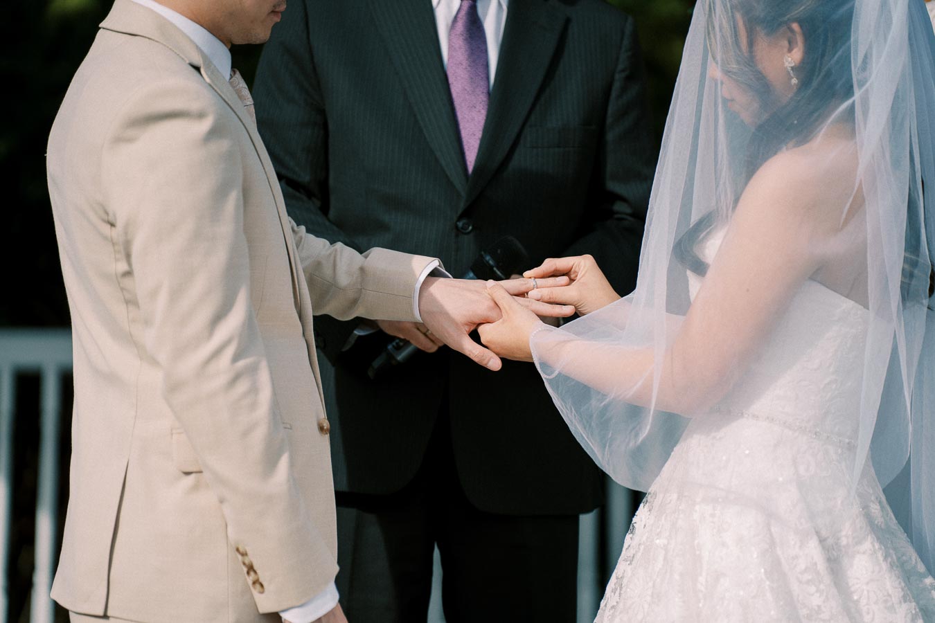 A bride and groom exchanging rings during their outdoor wedding ceremony, officiated by a person in a suit.