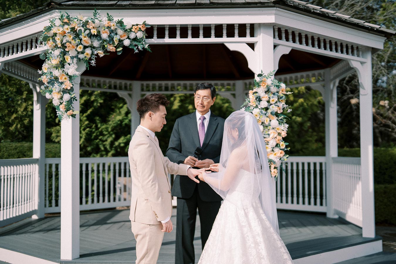 A bride and groom exchange rings during an outdoor wedding ceremony under a gazebo decorated with lush floral arrangements, as an officiant presides over the vows.