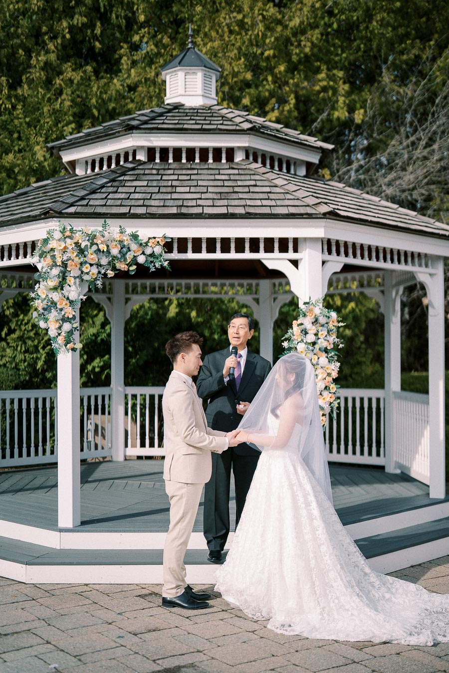 Wedding ceremony in a garden gazebo with a couple holding hands, officiant speaking, and floral decorations on a sunny day.