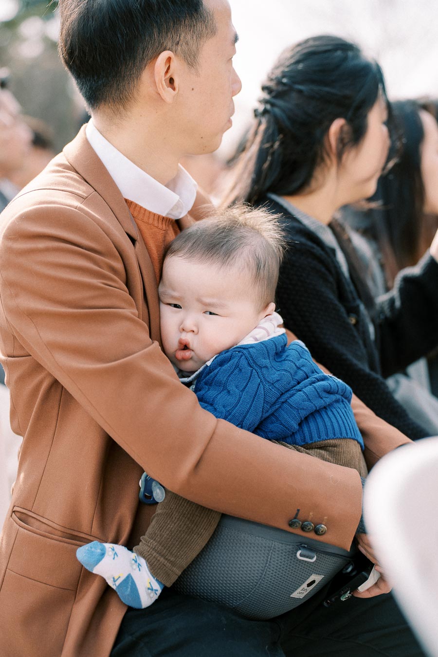 Father holding a baby in a blue sweater at an outdoor event, surrounded by other people.