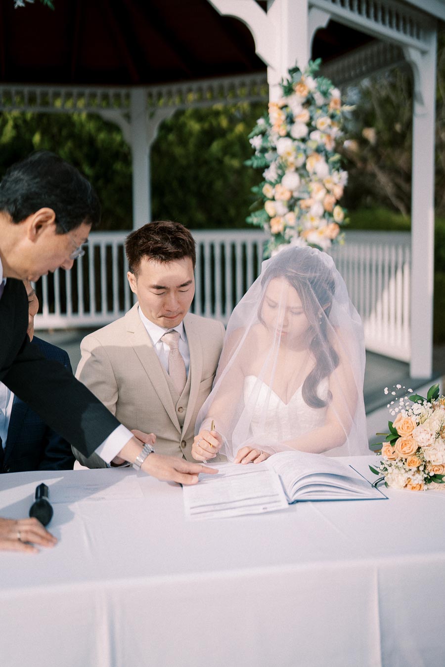 A bride and groom signing their wedding certificate under a decorated gazebo, with a bouquet of peach and white flowers on the table.