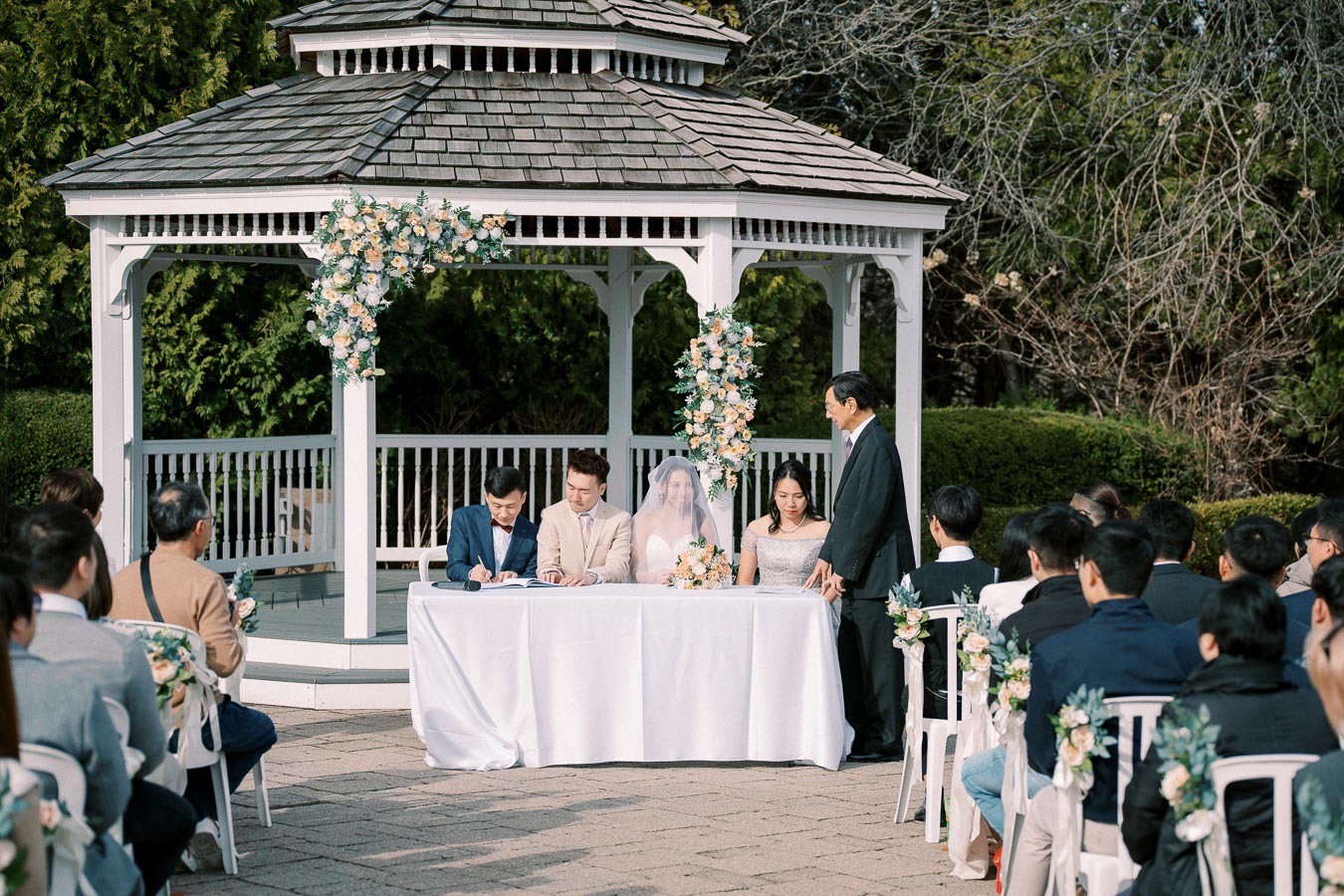 Outdoor wedding ceremony under a decorated gazebo, featuring a bride and groom seated at a table with witnesses signing documents, surrounded by guests.