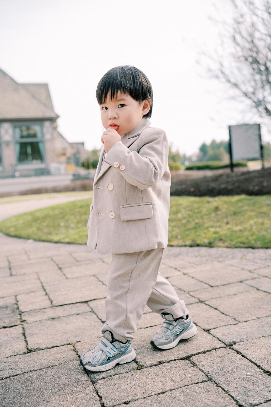 Young child wearing a beige suit and sneakers, standing on a paved path outside a building, holding a lollipop with a thoughtful expression.