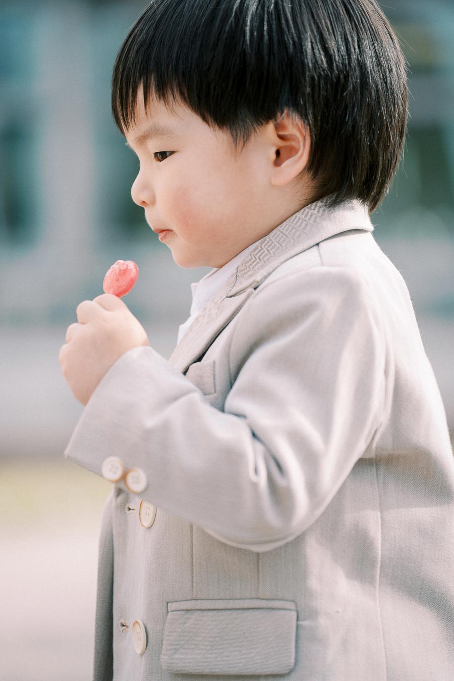 A young child in a beige suit holding a red lollipop outdoors, looking thoughtful.