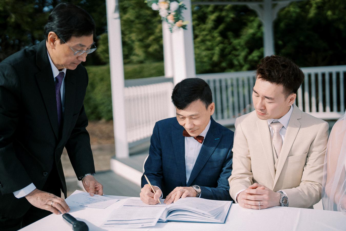 A group of men dressed in formal suits sign documents at an outdoor wedding ceremony, under a decorated gazebo surrounded by greenery.