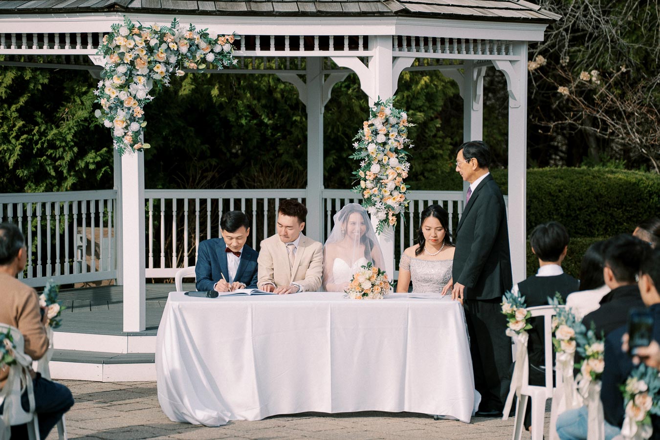 Outdoor wedding ceremony with a bride and groom seated at a decorated gazebo, surrounded by guests, as the groom signs the marriage certificate.