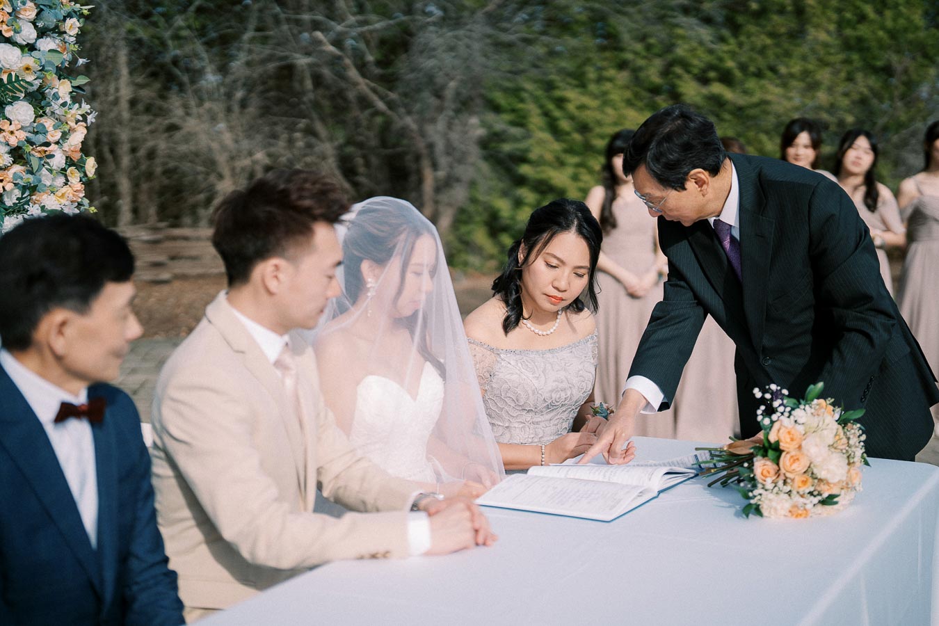 Wedding ceremony signing with bride, groom, and guests at an outdoor venue. A bouquet of flowers is on the table, enhancing the romantic setting.
