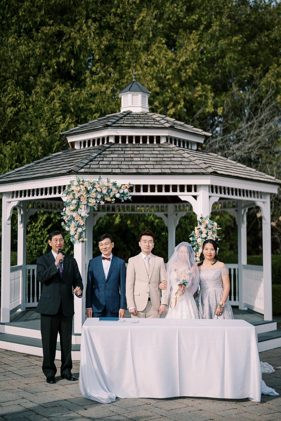 A wedding ceremony at an outdoor gazebo featuring a married couple and their family posing together. The scene shows a beautifully decorated gazebo with floral arrangements, set in a lush green garden, captured on a sunny day.