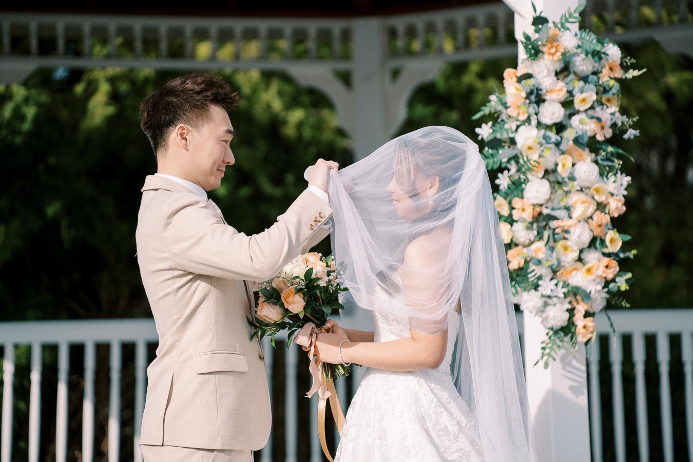 Man in beige suit lifting bride's veil during outdoor wedding ceremony with floral arch in background.