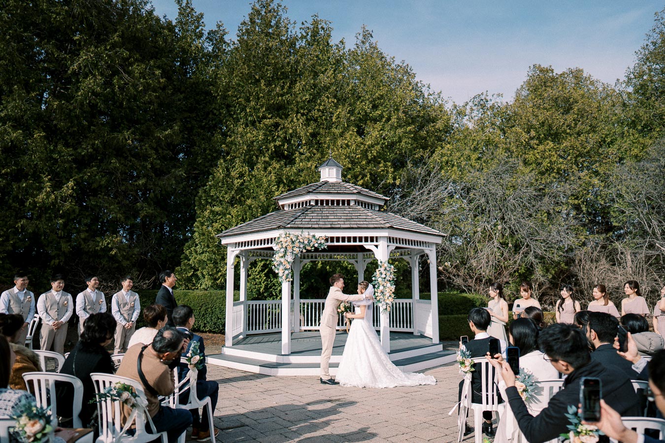 Outdoor wedding ceremony at a gazebo, featuring a bride in a white gown and a groom in a light suit, surrounded by guests seated on white chairs with flowers and greenery in the background.