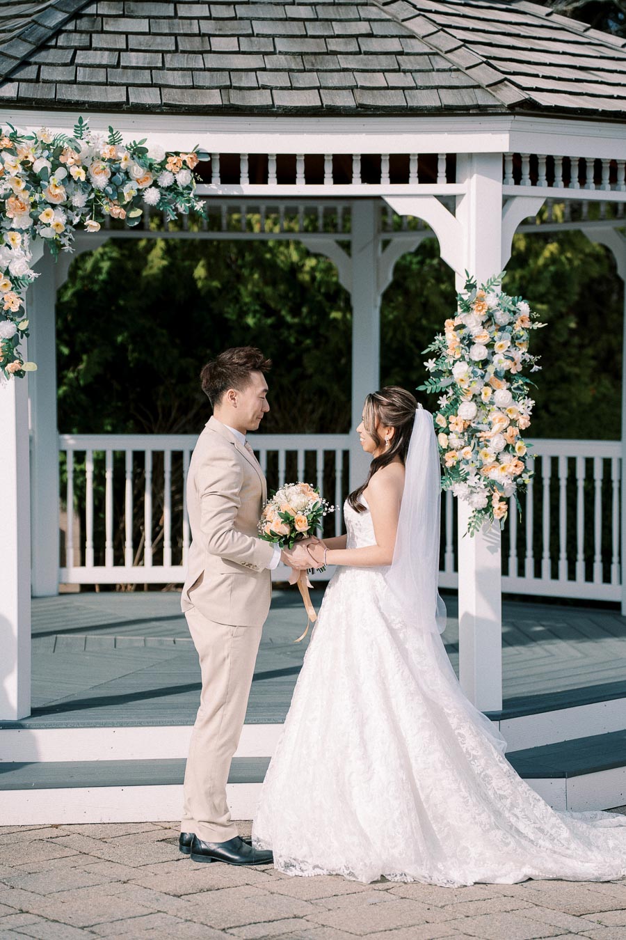 A couple in wedding attire exchanging vows in front of a beautifully decorated gazebo. The bride holds a bouquet of peach and white flowers, while the groom is dressed in a beige suit. The floral arrangements and serene outdoor setting enhance the romantic ambiance.