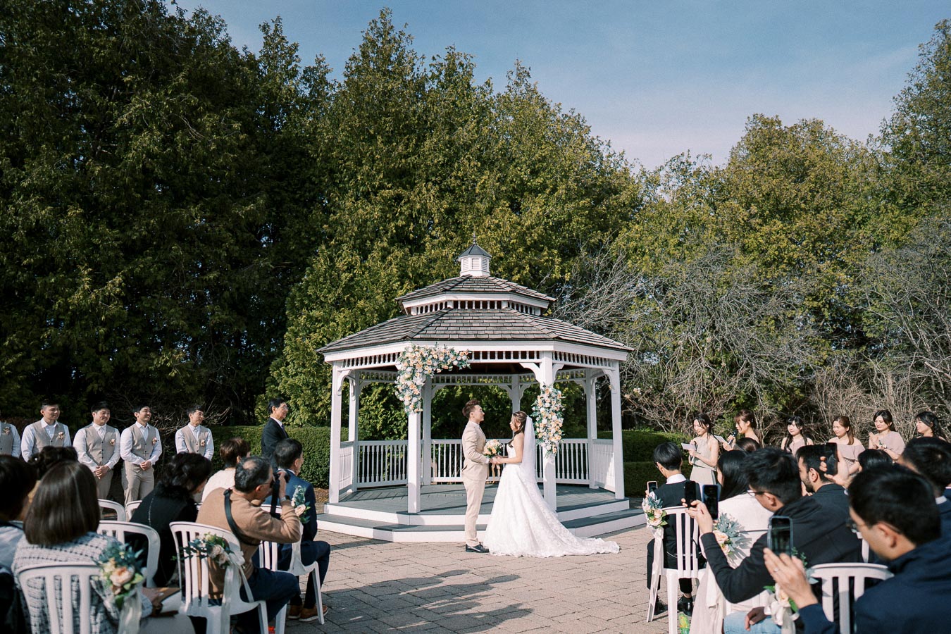 Outdoor wedding ceremony at a picturesque gazebo, with the bride and groom exchanging vows, surrounded by guests and lush greenery.