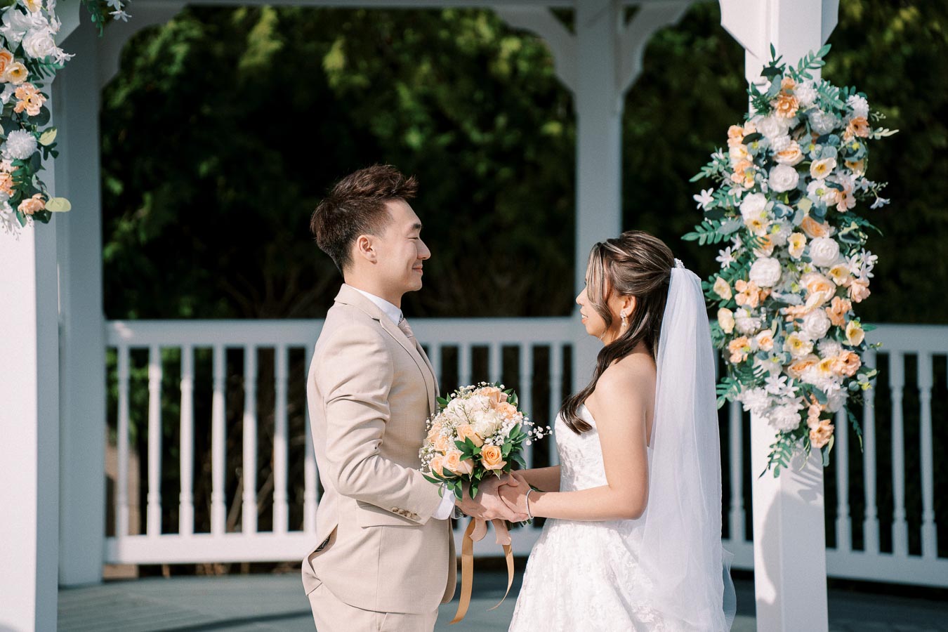 A bride and groom exchanging vows under a decorated gazebo; the groom in a beige suit holds a bouquet, and the bride in a white gown with a veil stands opposite, surrounded by floral arrangements.