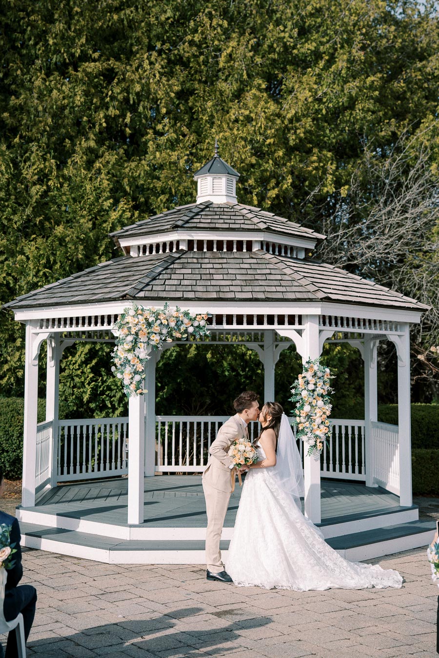 Outdoor wedding ceremony in front of a charming white gazebo, featuring a couple sharing a kiss. The bride wears a flowing white gown and holds a floral bouquet, while the groom is in a light tan suit. Elegant floral arrangements decorate the gazebo, set against a backdrop of lush greenery.
