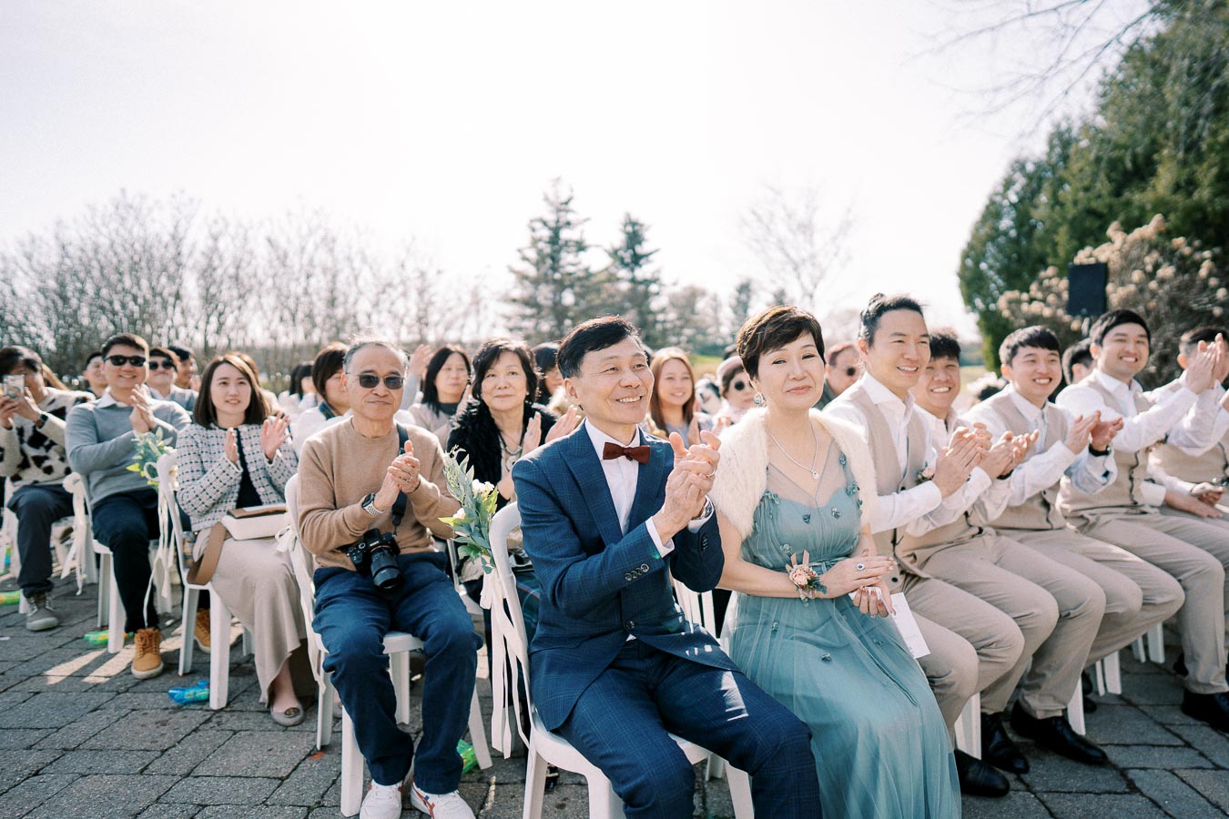 Outdoor wedding ceremony with seated guests applauding, featuring a well-dressed couple in formal attire, surrounded by friends and family, under a clear sky and natural backdrop.