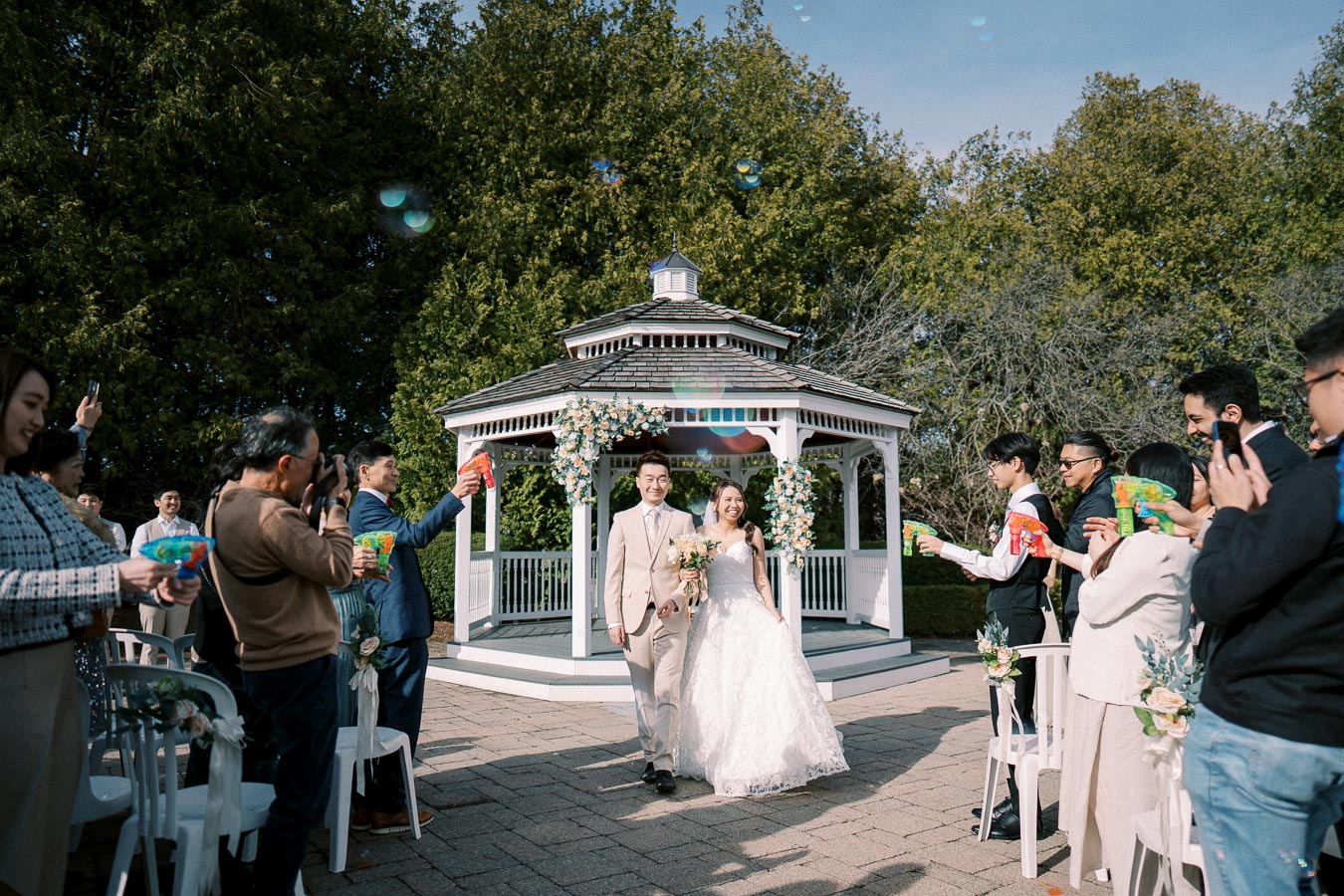 A newlywed couple happily walking down the aisle after their outdoor wedding ceremony, surrounded by guests blowing bubbles in celebration, near a beautifully decorated gazebo with greenery and flowers.