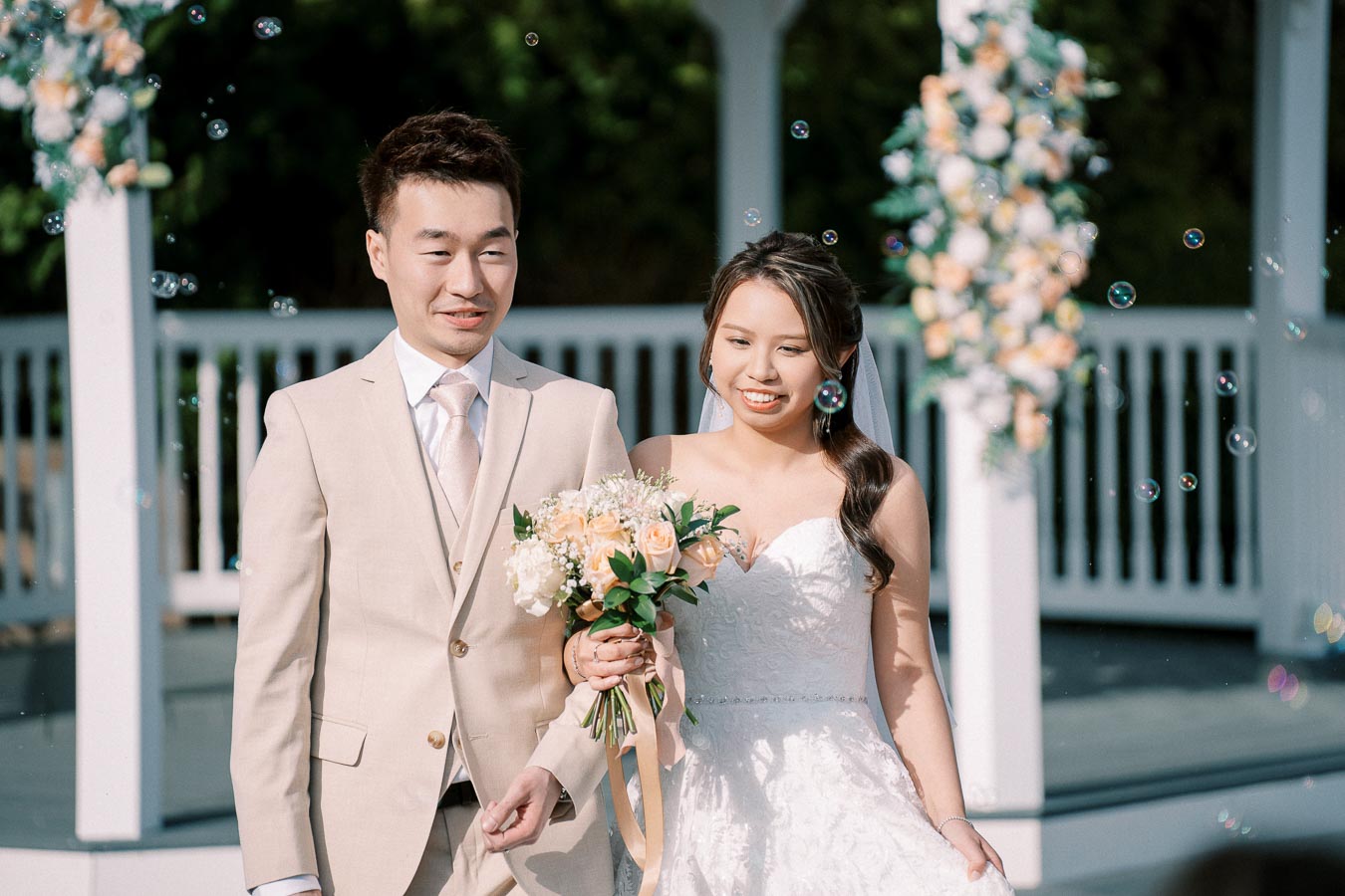 A happy bride and groom celebrating their wedding day, walking hand in hand under a gazebo adorned with floral decorations, with bubbles floating in the air.