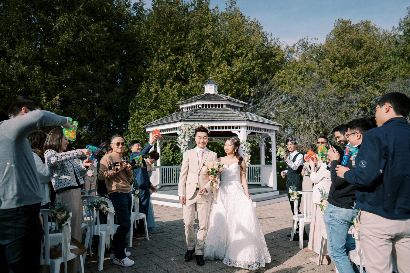 Bride and groom joyfully walking down the aisle under a decorated gazebo as guests blow bubbles at an outdoor wedding ceremony.