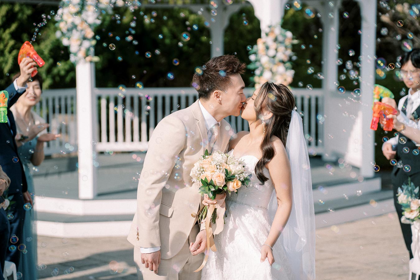 A bride and groom share a kiss at an outdoor wedding ceremony, surrounded by bubbles and guests celebrating with bubble guns. The bride holds a bouquet of peach and white flowers, and the groom is dressed in a light tan suit. The scene is joyful and romantic.