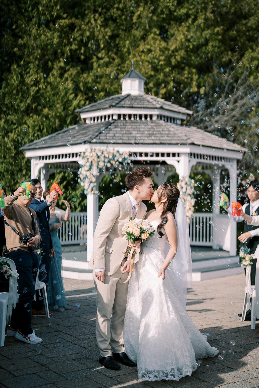 A newlywed couple shares a kiss in front of a decorated gazebo surrounded by trees, as guests celebrate by blowing bubbles at their outdoor wedding ceremony. The bride holds a bouquet of flowers, and the groom wears a light-colored suit.