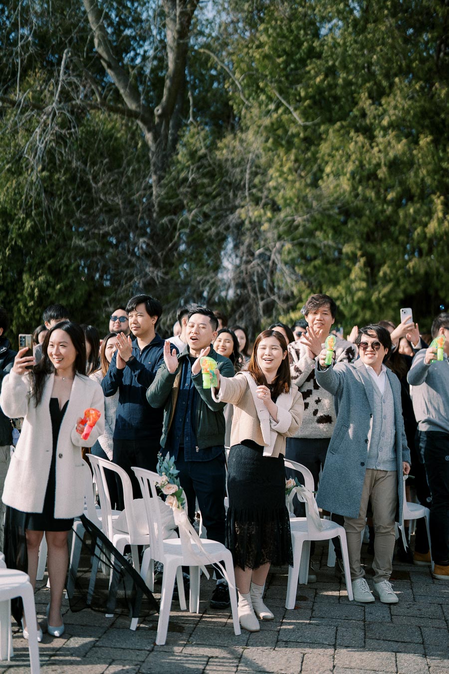 A group of people smiling and clapping while holding colorful party poppers at an outdoor event, with green trees in the background.