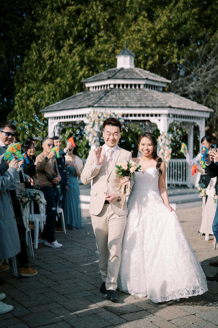 Bride and groom joyfully walking down the aisle in a picturesque outdoor wedding ceremony, surrounded by guests blowing bubbles, with a charming gazebo adorned with flowers in the background.