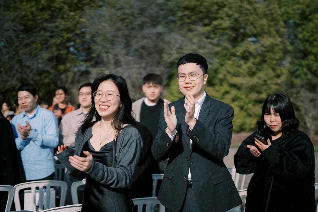 Group of people outdoors clapping and smiling during an event, with trees in the background.