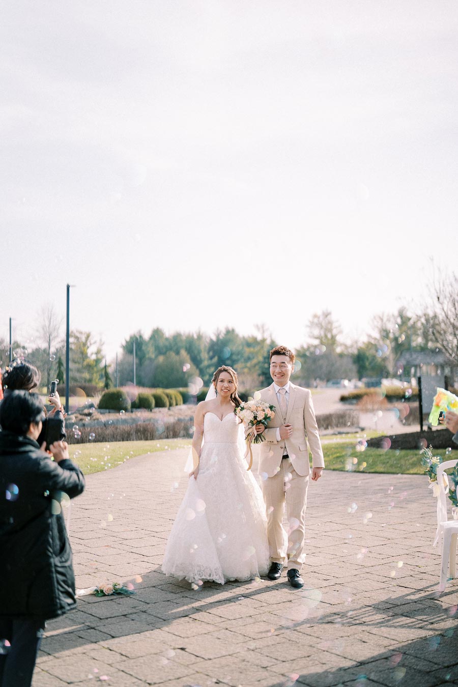 A bride and groom happily walk down an outdoor aisle on a sunny day, surrounded by guests taking photos and blowing bubbles, with a scenic garden backdrop.