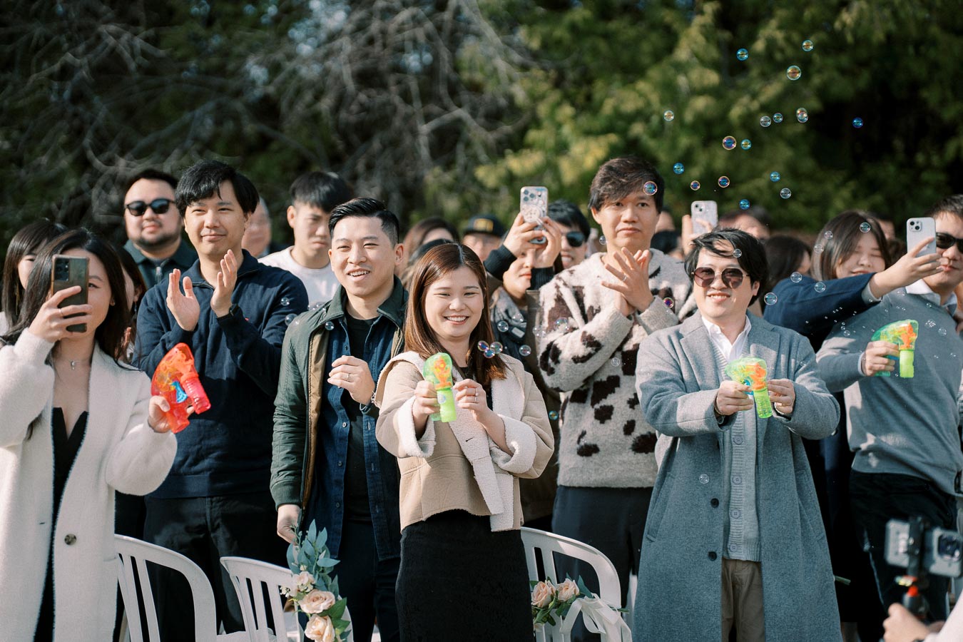 A joyful crowd at an outdoor event celebrating with bubble guns, smartphones capturing the moment; people smiling and clapping.