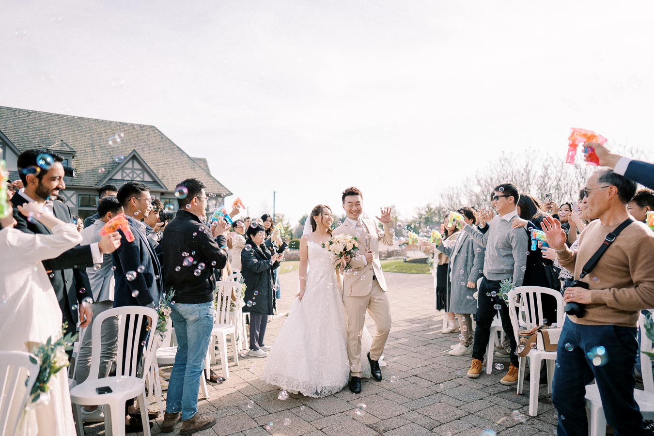 Bride and groom happily walking down an outdoor aisle surrounded by wedding guests blowing bubbles, capturing a joyful celebration.