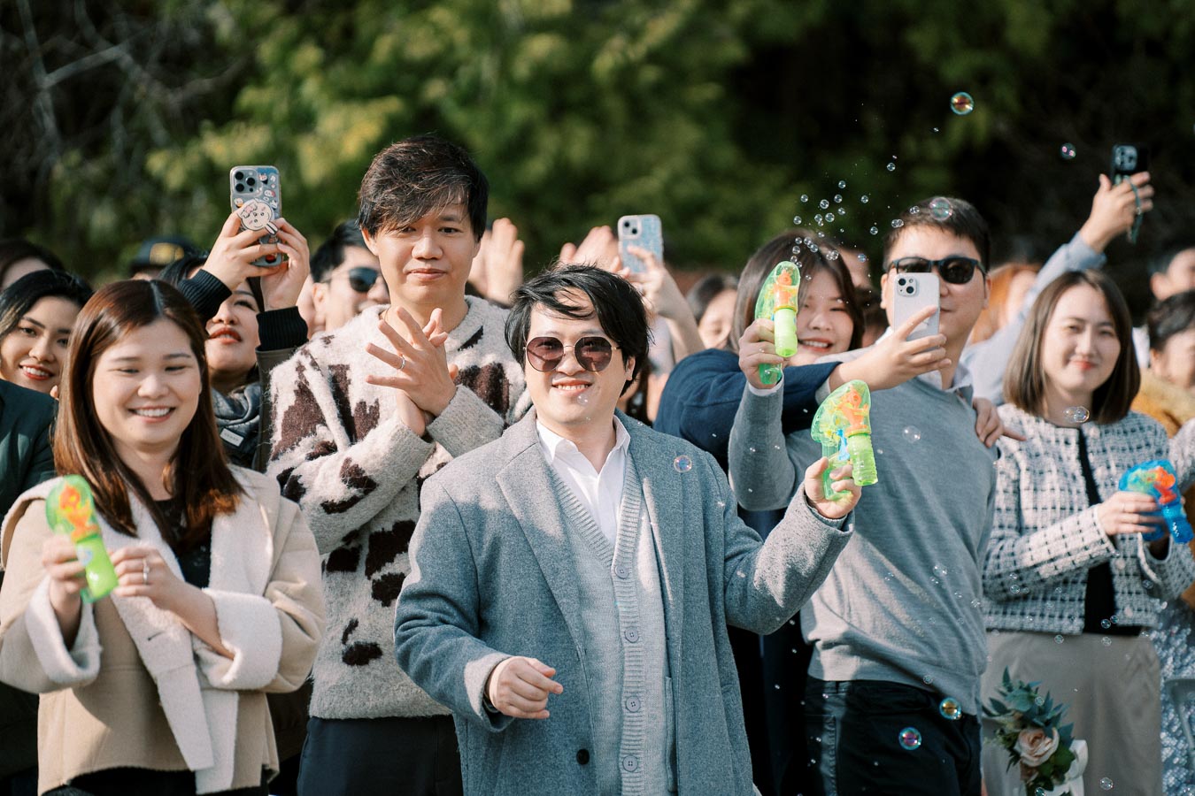 Group of people outdoors celebrating with bubble guns, smiling and taking photos, with greenery in the background.