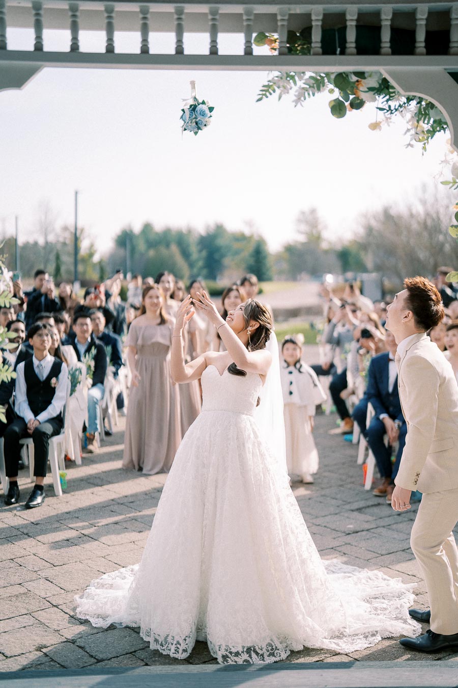 Bride in a white wedding gown tossing bouquet with guests watching at outdoor ceremony on a sunny day