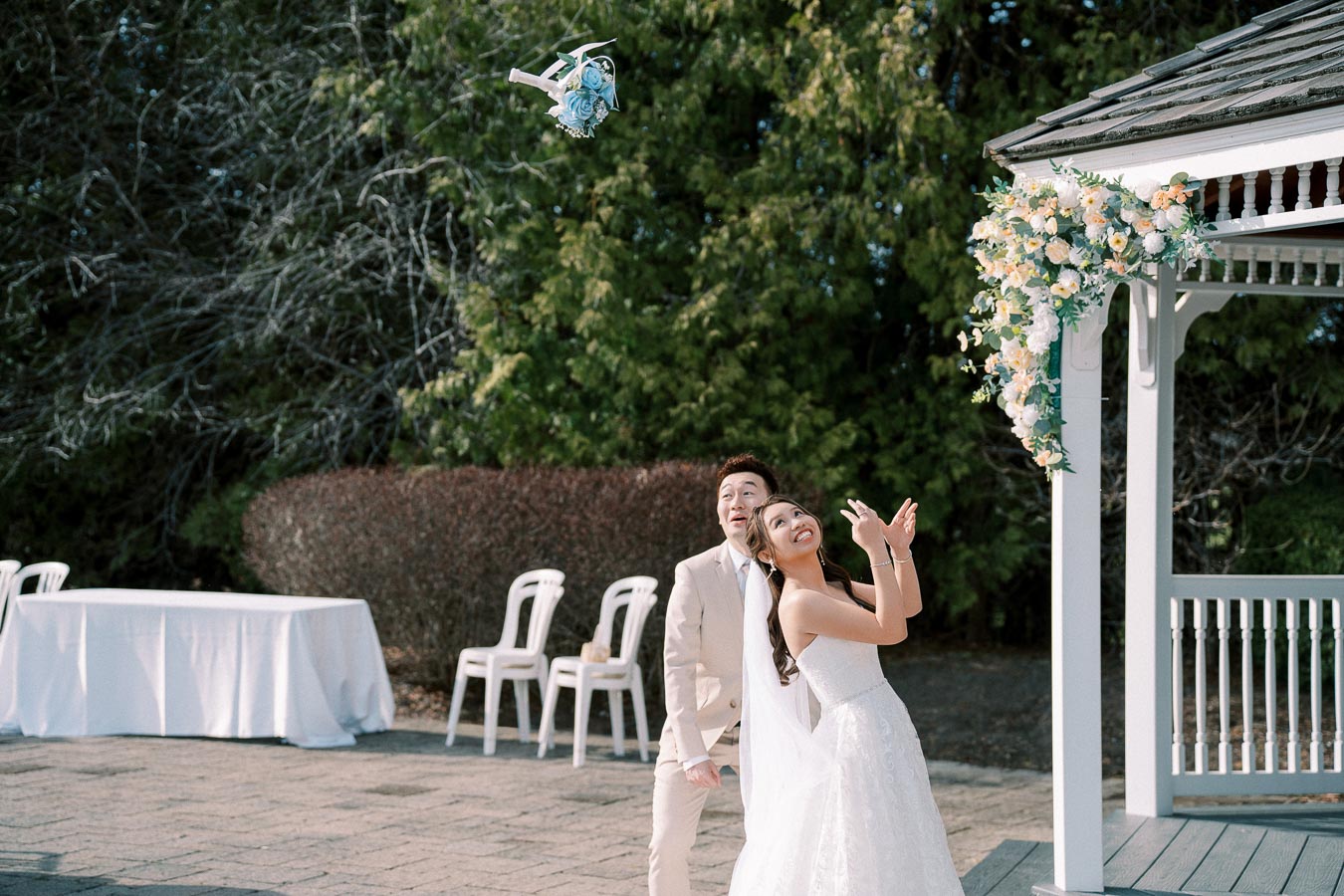 Bride and groom joyfully reacting to a drone capturing their wedding, standing near a gazebo adorned with floral decorations.