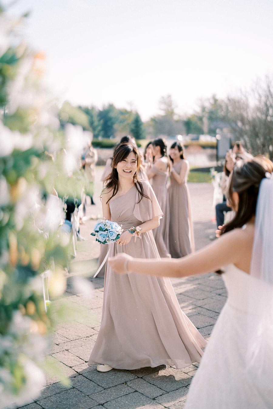 Bridesmaid in taupe dress holding bouquet of blue flowers, smiling at outdoor wedding ceremony with blurred bride in foreground.