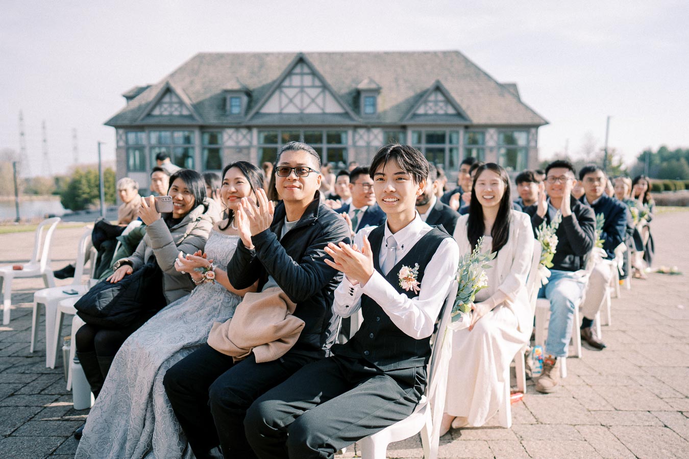 Outdoor wedding ceremony guests sitting and clapping, with a scenic building in the background.