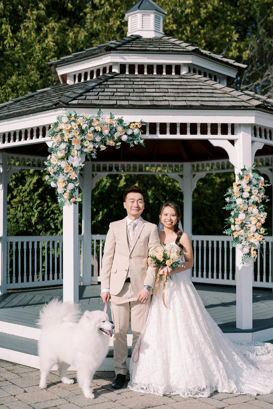 A joyful couple in wedding attire poses with their fluffy white dog in front of a beautifully decorated gazebo with floral arrangements.
