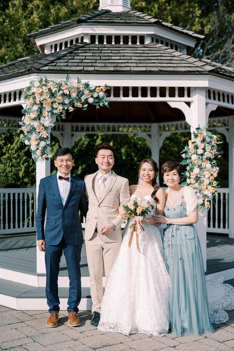 A newlywed couple poses happily with their parents in front of a beautifully decorated gazebo, adorned with white and peach flowers, under the bright sunlight, highlighting a charming outdoor wedding setting.