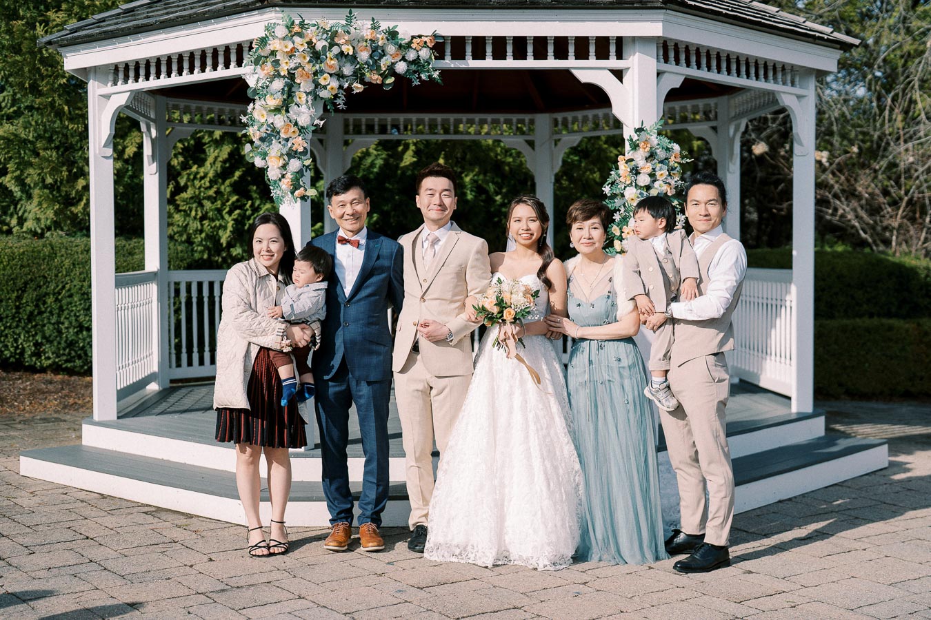 A joyful wedding celebration with a bride in a white gown and groom in a beige suit surrounded by family members, standing in front of a beautifully decorated gazebo adorned with flowers.